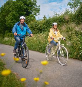 Two adult cyclists wearing helmets ride side by side along a peaceful Greenway path, surrounded by greenery and wildflowers on a relaxing countryside cycle.