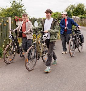 Three teenagers walking with their bicycles along a quiet Greenway lane, chatting and enjoying a relaxed cycling outing together.