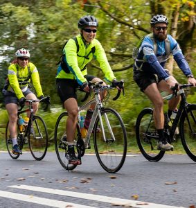 Three adult cyclists wearing helmets and high-visibility cycling gear ride together along a tree-lined Greenway route, enjoying an energetic group ride through the countryside.