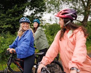 Three women wearing helmets and waterproof jackets cycle together along the Greenway, smiling and chatting as they enjoy a social outdoor bike ride.