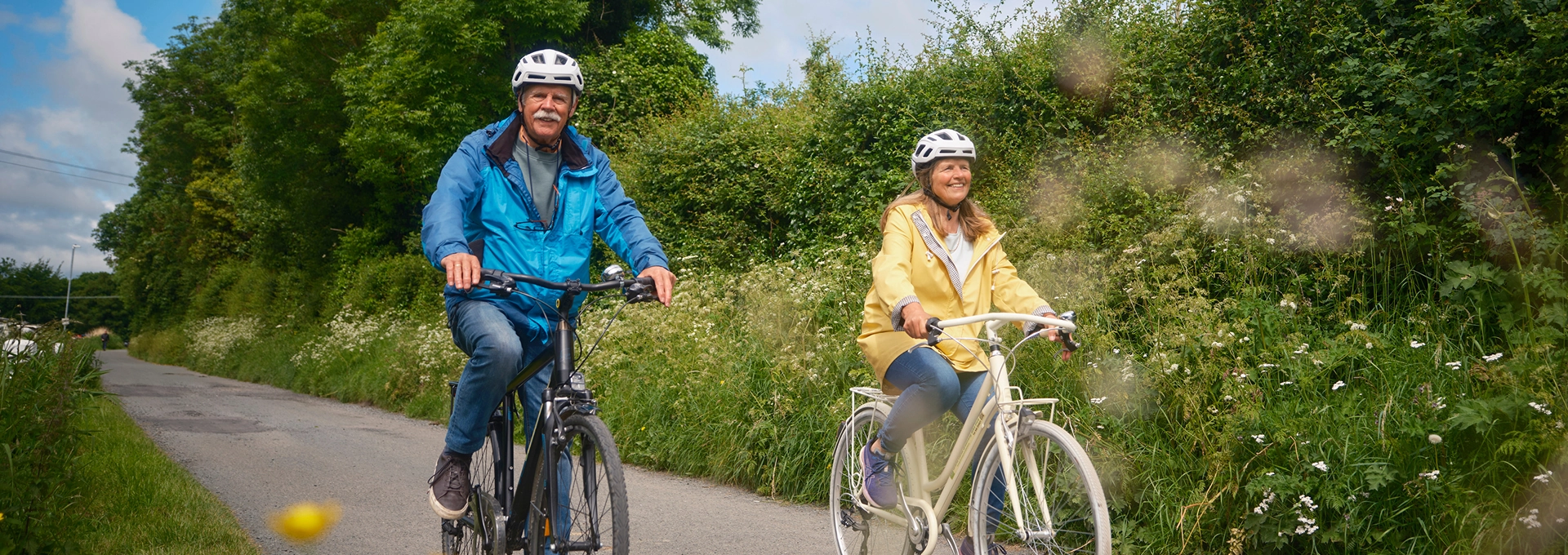 Two adult cyclists wearing helmets ride side by side along a peaceful Greenway path, surrounded by greenery and wildflowers on a relaxing countryside cycle.
