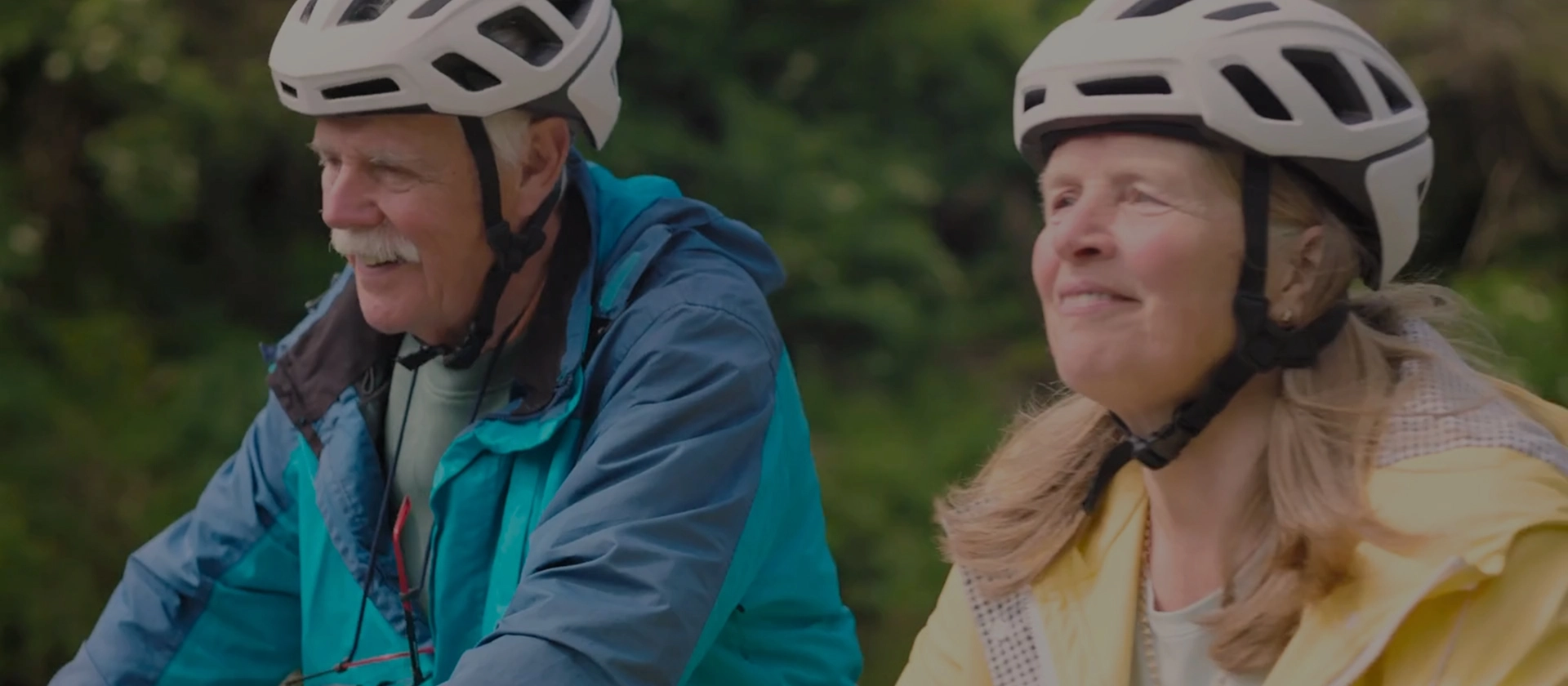 An older couple wearing helmets enjoy a leisurely bike ride together along the Greenway, smiling as they take in the peaceful surroundings.