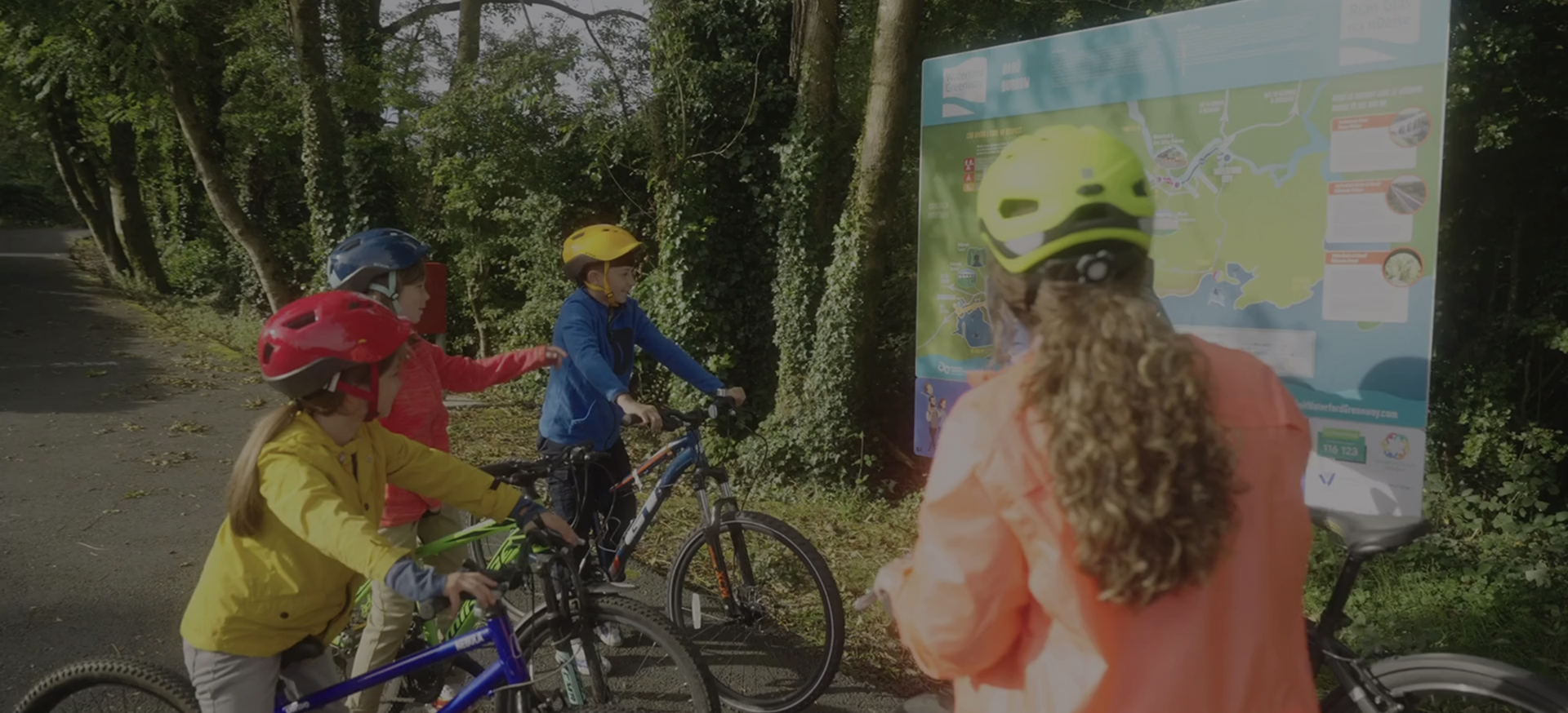 A group of children wearing helmets stop along the Greenway to look at a route map, learning about the trail and planning their cycling journey together.