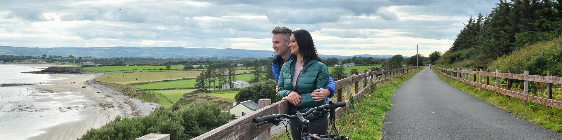A couple stands beside their bicycles on the Waterford Greenway, pausing to admire the scenic coastal views and countryside stretching into the distance.