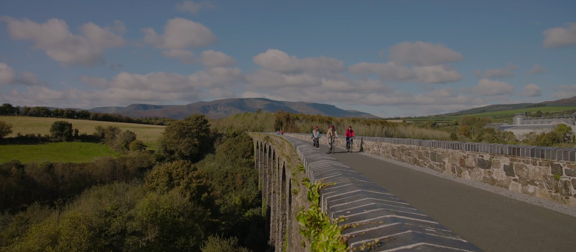 Cyclists ride across a historic stone viaduct on the Waterford Greenway, surrounded by panoramic views of countryside and distant mountains.