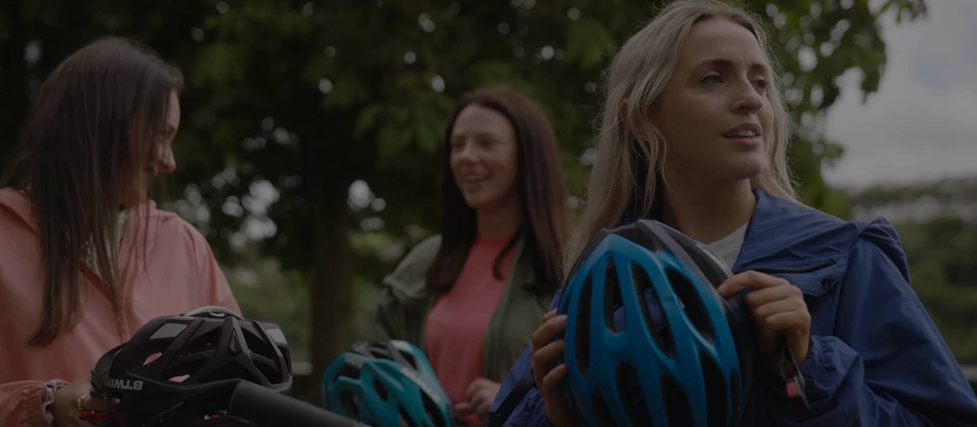 Three women holding cycling helmets chat and smile together outdoors, preparing for a Greenway bike ride through the countryside.